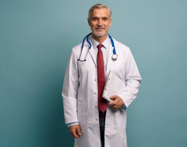 Steteskoplu, olgun bir tıp uzmanı. Kameraya gülümsüyor, elinde notlarla kâğıtlar tutuyor. Mavi arka planda izole edilmiş.