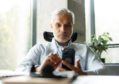 Orta yaşlı iş adamı elinde akıllı telefon tutarak çalışıyor. Olgun 45 yaşındaki işadamı yöneticisi, iş sahibi veya girişimci cep telefonu kullanarak masada oturuyor.