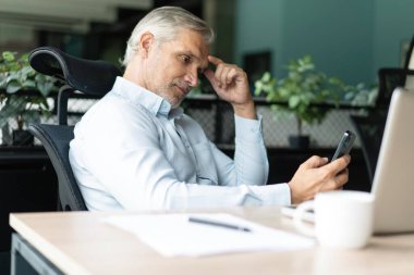 Orta yaşlı iş adamı elinde akıllı telefon tutarak çalışıyor. Olgun 45 yaşındaki işadamı yöneticisi, iş sahibi veya girişimci cep telefonu kullanarak masada oturuyor.