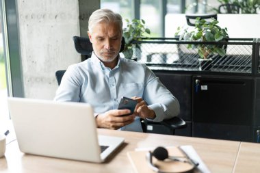 Orta yaşlı iş adamı elinde akıllı telefon tutarak çalışıyor. Olgun 45 yaşındaki işadamı yöneticisi, iş sahibi veya girişimci cep telefonu kullanarak masada oturuyor.