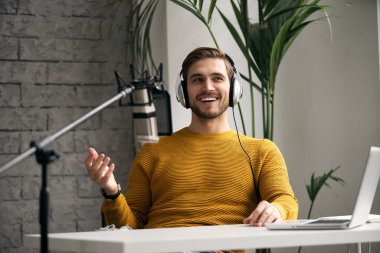 Adam podcaster etkili blogcu gülümseyerek stüdyoda kulaklık, dizüstü bilgisayar ve kulaklık kullanarak canlı yayın yapıyor. Erkek radyo sunucusu podcast veya röportaj yapıyor