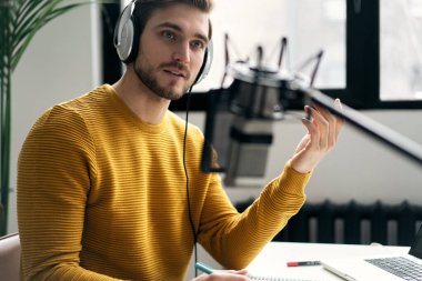 Adam podcaster etkili blogcu gülümseyerek stüdyoda kulaklık, dizüstü bilgisayar ve kulaklık kullanarak canlı yayın yapıyor. Erkek radyo sunucusu podcast veya röportaj yapıyor