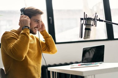 Adam podcaster etkili blogcu gülümseyerek stüdyoda kulaklık, dizüstü bilgisayar ve kulaklık kullanarak canlı yayın yapıyor. Erkek radyo sunucusu podcast veya röportaj yapıyor
