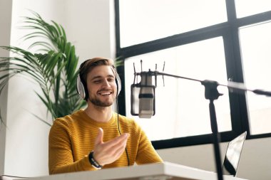 Adam podcaster etkili blogcu gülümseyerek stüdyoda kulaklık, dizüstü bilgisayar ve kulaklık kullanarak canlı yayın yapıyor. Erkek radyo sunucusu podcast veya röportaj yapıyor