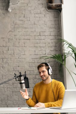 Adam podcaster etkili blogcu gülümseyerek stüdyoda kulaklık, dizüstü bilgisayar ve kulaklık kullanarak canlı yayın yapıyor. Erkek radyo sunucusu podcast veya röportaj yapıyor
