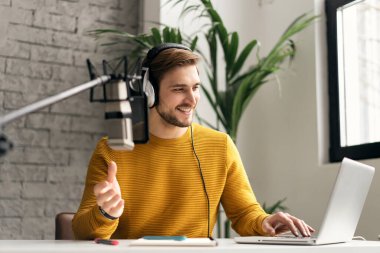Adam podcaster etkili blogcu gülümseyerek stüdyoda kulaklık, dizüstü bilgisayar ve kulaklık kullanarak canlı yayın yapıyor. Erkek radyo sunucusu podcast veya röportaj yapıyor