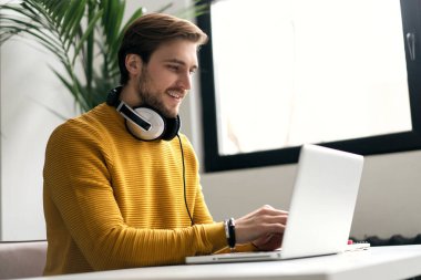 Casually dressed young designer listening music on headphones while working on a laptop at a desk in an office