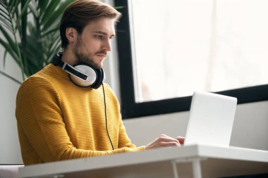 Ofisteki bir masada dizüstü bilgisayarla çalışırken kulaklıkla müzik dinleyen sıradan giyinmiş genç bir tasarımcı.