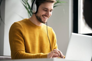 Casually dressed young designer listening music on headphones while working on a laptop at a desk in an office