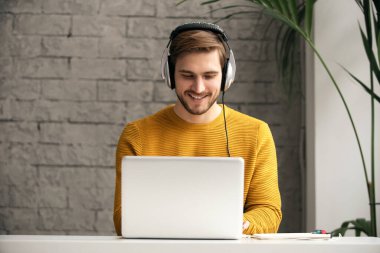 Ofisteki bir masada dizüstü bilgisayarla çalışırken kulaklıkla müzik dinleyen sıradan giyinmiş genç bir tasarımcı.