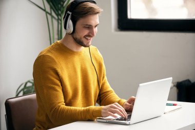 Ofisteki bir masada dizüstü bilgisayarla çalışırken kulaklıkla müzik dinleyen sıradan giyinmiş genç bir tasarımcı.
