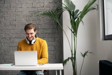 Ofisteki bir masada dizüstü bilgisayarla çalışırken kulaklıkla müzik dinleyen sıradan giyinmiş genç bir tasarımcı.