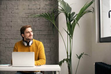 Ofisteki bir masada dizüstü bilgisayarla çalışırken kulaklıkla müzik dinleyen sıradan giyinmiş genç bir tasarımcı.