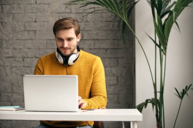 Casually dressed young designer listening music on headphones while working on a laptop at a desk in an office