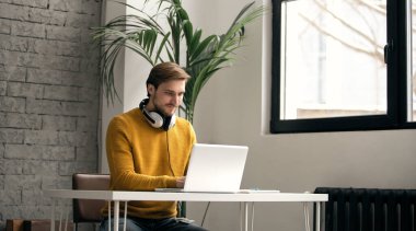 Ofisteki bir masada dizüstü bilgisayarla çalışırken kulaklıkla müzik dinleyen sıradan giyinmiş genç bir tasarımcı.