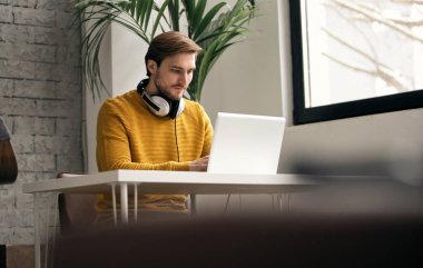 Ofisteki bir masada dizüstü bilgisayarla çalışırken kulaklıkla müzik dinleyen sıradan giyinmiş genç bir tasarımcı.