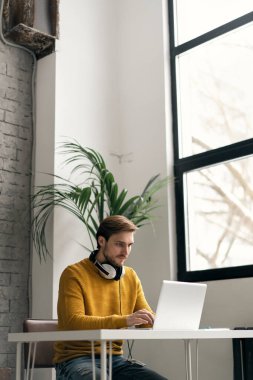 Ofisteki bir masada dizüstü bilgisayarla çalışırken kulaklıkla müzik dinleyen sıradan giyinmiş genç bir tasarımcı.