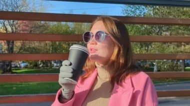 Paltolu genç bayanın yürüdüğü, elinde sıcak kahveyle kağıt bardak tuttuğu yakın plan. Çekici bir kadın sıcak kahve içiyor, kokuyor, aromanın tadını çıkarıyor, gülümsüyor. Mutluluk ve neşe kavramı. Yüksek