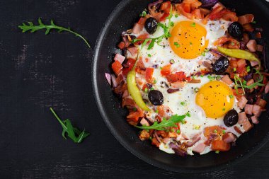 Fried eggs with vegetables on pan - pepperoni, tomatoes, ham, onion and black olives. Ketogenic breakfast. Top view, above