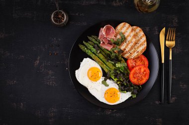 Breakfast. Fried egg, bread toast, green asparagus, tomatoes and jamon on black plate. Top view, above