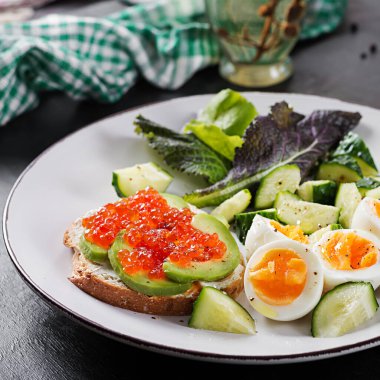 Breakfast. Healthy open sandwich on  toast with avocado and red caviar, boiled eggs, cucumber salad on white plate. Healthy protein food.
