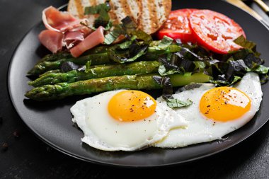 Breakfast. Fried egg, bread toast, green asparagus, tomatoes and jamon on black plate.
