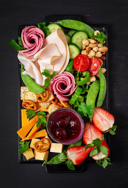 Antipasto platter cold sliced ham, salami, crackers, strawberries, vegetables and cheese platter on  board over dark background. Appetizers table with italian antipasti snacks. Top view