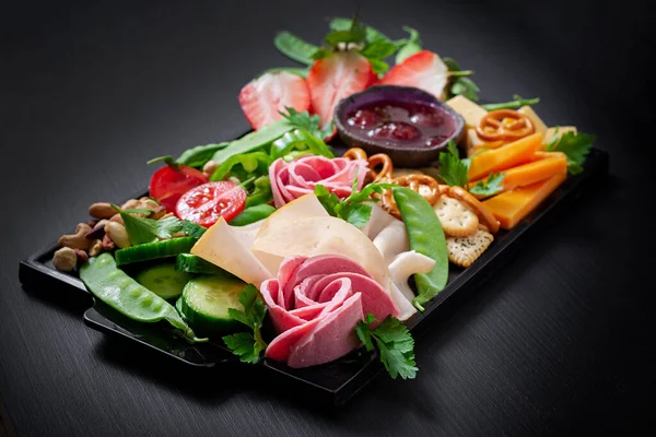 Antipasto platter cold sliced ham, salami, crackers, strawberries, vegetables and cheese platter on  board over dark background. Appetizers table with italian antipasti snacks.