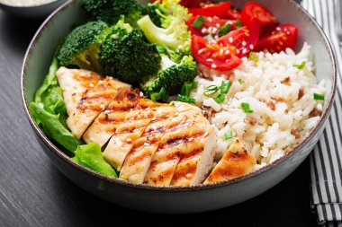 Delicious buddha bowl with grilled chicken, fresh vegetables and rice on a dark background.
