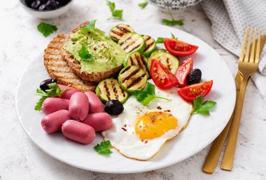 Fried egg with mini sausage, tomatoes, grilled zucchini, black olives and toasts with guacamole. English Breakfast. Brunch.