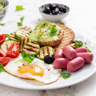 Fried egg with mini sausage, tomatoes, grilled zucchini, black olives and toasts with guacamole. English Breakfast. Brunch.