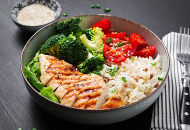 Delicious buddha bowl with grilled chicken, fresh vegetables and rice on a dark background.