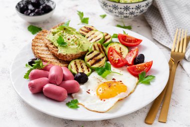 Fried egg with mini sausage, tomatoes, grilled zucchini, black olives and toasts with guacamole. English Breakfast. Brunch.