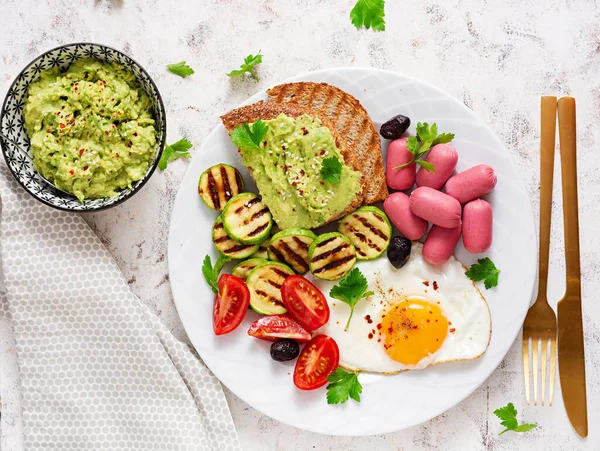 Fried egg with mini sausage, tomatoes, grilled zucchini, black olives and toasts with guacamole. English Breakfast. Brunch. Top view