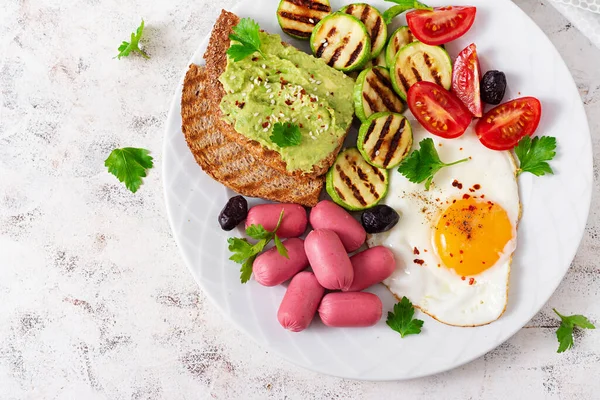 Fried egg with mini sausage, tomatoes, grilled zucchini, black olives and toasts with guacamole. English Breakfast. Brunch. Top view