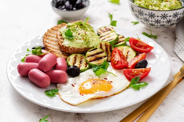 Fried egg with mini sausage, tomatoes, grilled zucchini, black olives and toasts with guacamole. English Breakfast. Brunch.