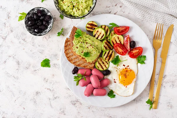 Fried egg with mini sausage, tomatoes, grilled zucchini, black olives and toasts with guacamole. English Breakfast. Brunch. Top view
