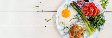 Ketojenik kahvaltı. Kahvaltıda. Kızarmış yumurta, somonlu gevrek somun, yeşil kuşkonmaz, taze domates ve beyaz tabakta kırmızı soğan. Üst görünüm, afiş