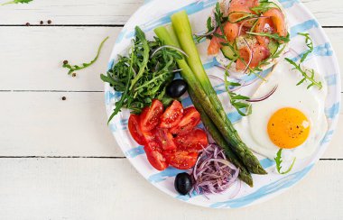 Ketojenik kahvaltı. Kahvaltıda. Kızarmış yumurta, somonlu gevrek somun, yeşil kuşkonmaz, taze domates ve beyaz tabakta kırmızı soğan. Üst manzara, düz uzanma