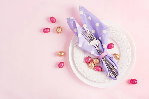 イースターテーブルの設定 ピンクの背景にウサギ イースター チョコレートの卵の形で折り畳まれたナプキンとホワイトプレート ハッピーイースターの休日の概念 トップビュー フラットレイアウト — ストック写真