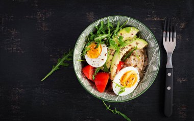 Kahvaltıda haşlanmış yumurta, avokado, domates ve yeşil otlu yulaf lapası. Sağlıklı dengeli yiyecekler. Üst manzara, düz uzanma