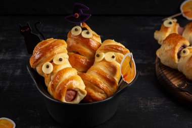 Halloween food. Scary sausage mummies in dough with funny eyes on dark background. Funny decoration.