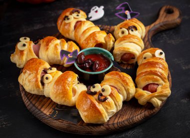 Cadılar Bayramı yemeği. Ürkütücü sosis mumyaları hamur içinde ve koyu arkaplanda komik gözleri var..