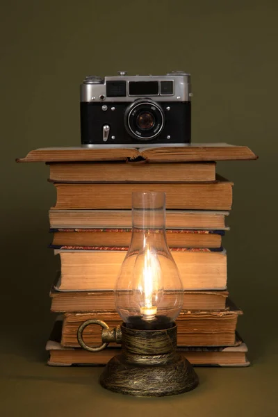 Old books and a hand lamp with bulb light, stylized as antique. There is an old camera on top of the books. Objects placed on an olive background.