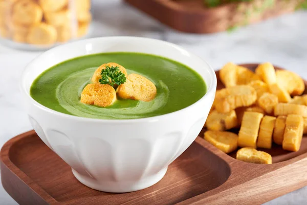 Traditional green broth or green soup with toast in the center and sour cream to decorate. wooden container with breadsticks on the side and ingredients in the background. on a marble stone.