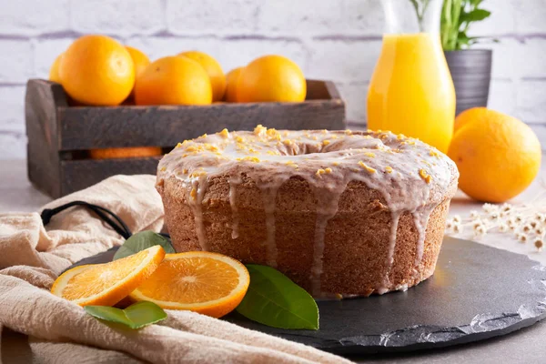 Traditional Brazilian orange cake with orange and lemon zest topping on a stone table and white brick rustic background. Oranges inside a rustic wooden crate. Orange juice and orange slices.