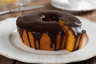 Homemade round carrot cake with chocolate coating, typical Brazilian food at a June party and a plate on a rustic wooden table with one slice.