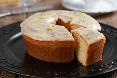 Homemade round lemon cake with topping and lemon zest on a plate on a rustic wooden table with on slice.