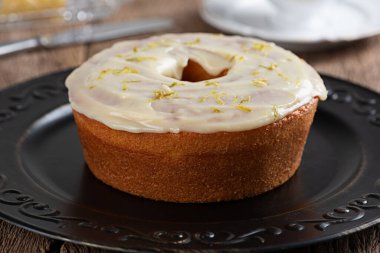 Homemade round lemon cake with topping and lemon zest on a plate on a rustic wooden table.