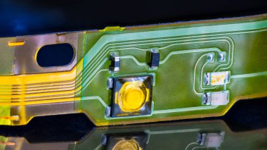 Close-up of electronic flex printed circuit with reflection on a shiny black background. Flat plastic strip with small surface mounted components on green-yellow flexible PCB from dismantled headphones. clipart